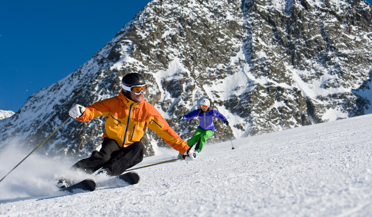 skiier in orange jacket skiing down a slope in front of another skier who is wearing purple.