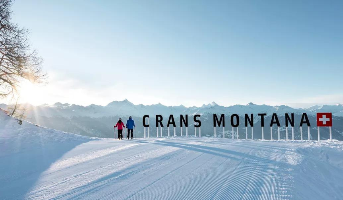 horizontal piste, with groomed snow and two skiers in the background overlooking the valley. There are letters on stilts reading: Crans Montana with the Swiss flag.