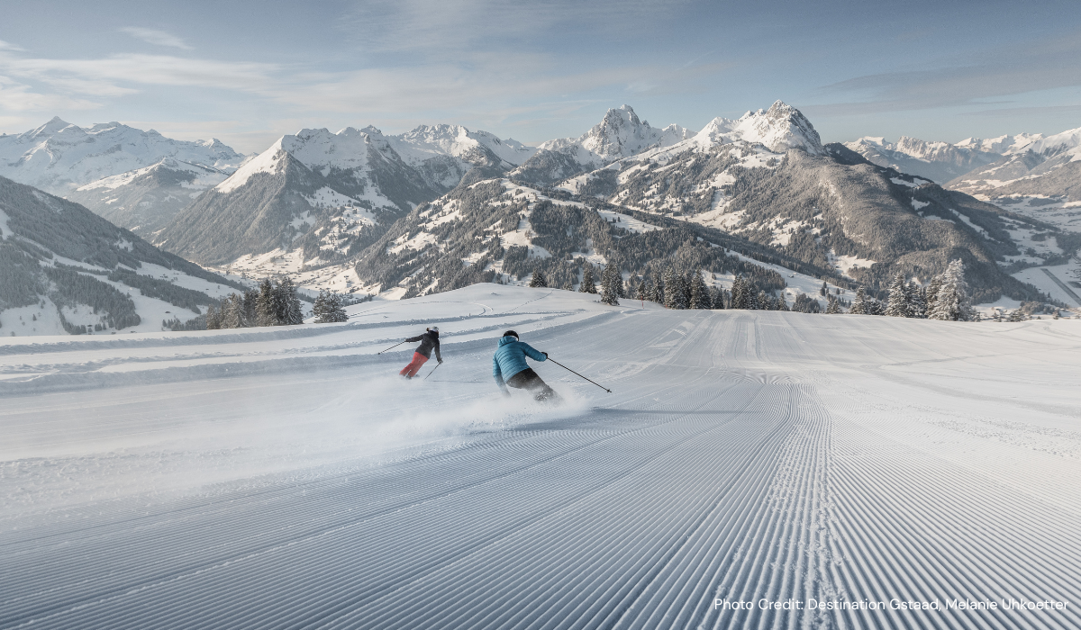 corduroy slopes with two skiers carving down the hill, mountains in the background.