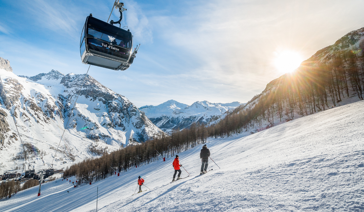 Slope side with setting sun in the background, three skiers on the piste and a black cable gondola over head,