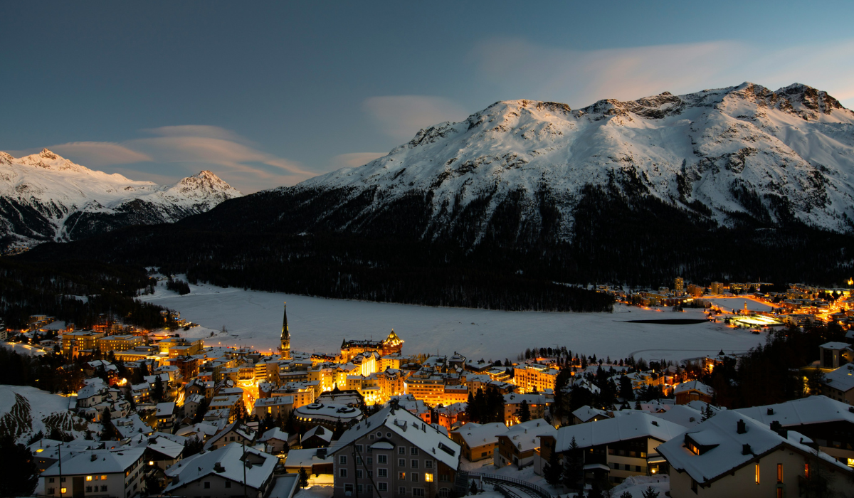 Twighlight hour looking over a snowy covered St Moritz village with the warm lights of the buildings and a frozen white lake.