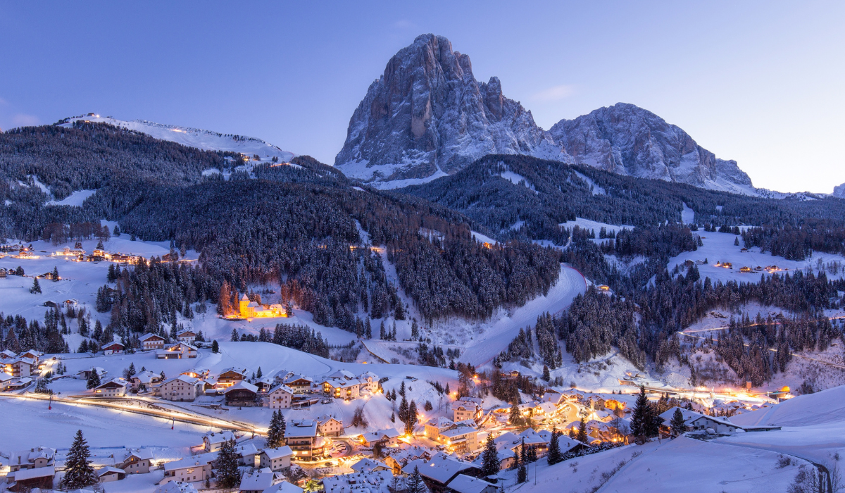 Iconic Dolomite peak in the background, with woodland and a twighlight hour with warm lighting in the buildings below in the valley.