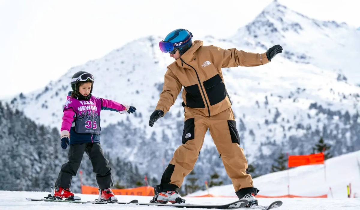 Ski Instructor teaching a child how to lean when skiing. They are both in ski-wear and have a snow covered mountain backdrop.