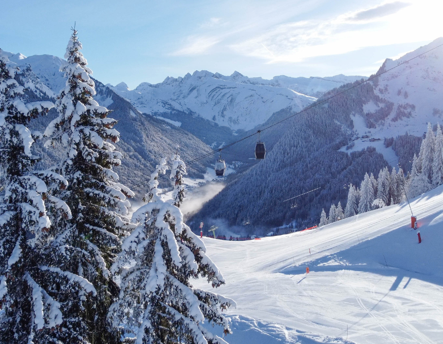 Stunning snowy vista of a sloping piste, cable car in background and snowy pine trees, blue sky and snow covered mountains.