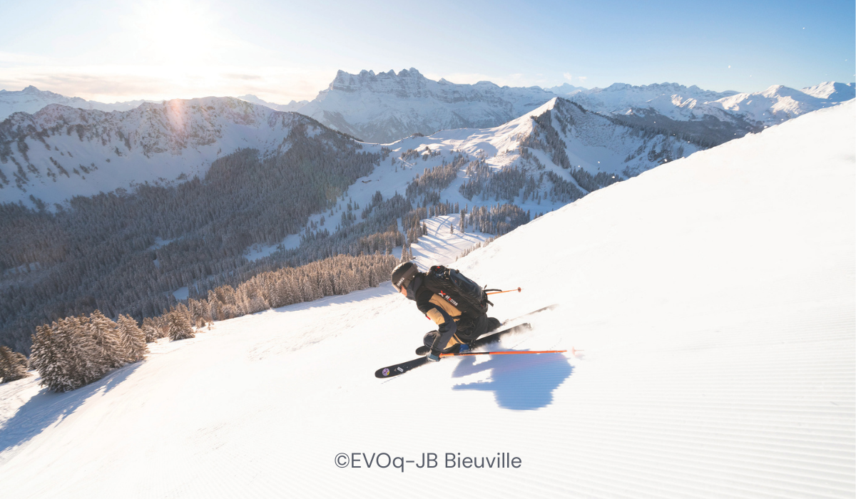 Skiier skiing down a well-groomed piste, the sun rising on the horizon and the mountains in the background covered in snow.