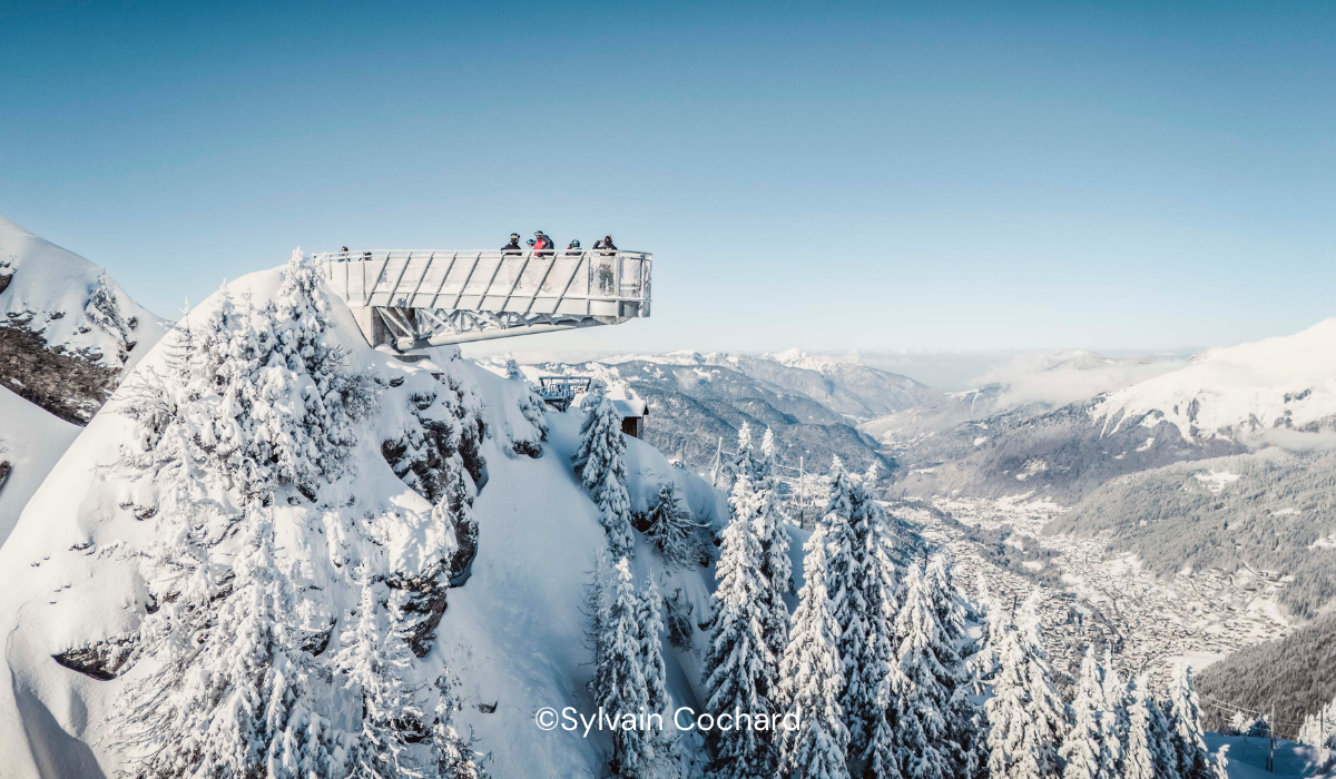 Platform suspended over a cliff top, with snow-covered pine trees and a valley with clear blue sky.