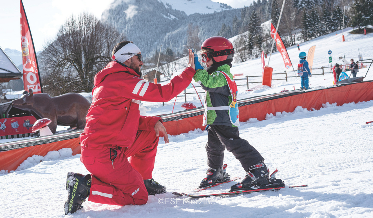 Ski instructor in ESF Red inform, crouching down to high-five a learner child.