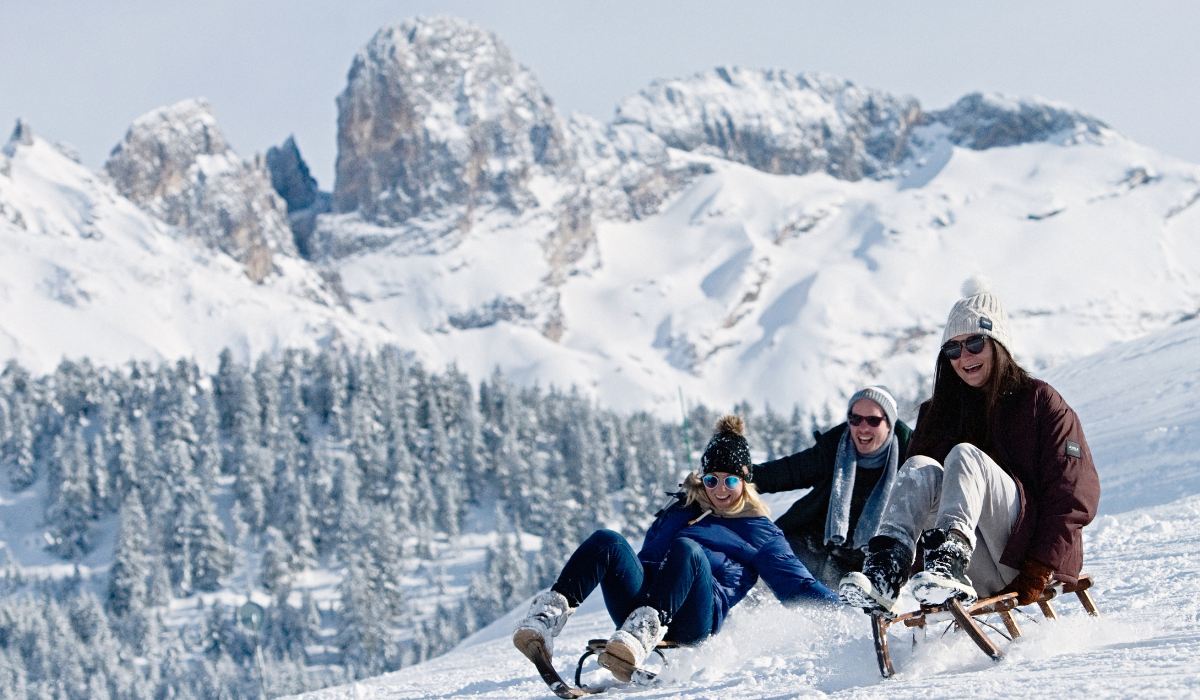 Three adults tabogganing down a slope with snowy peaks in the background.