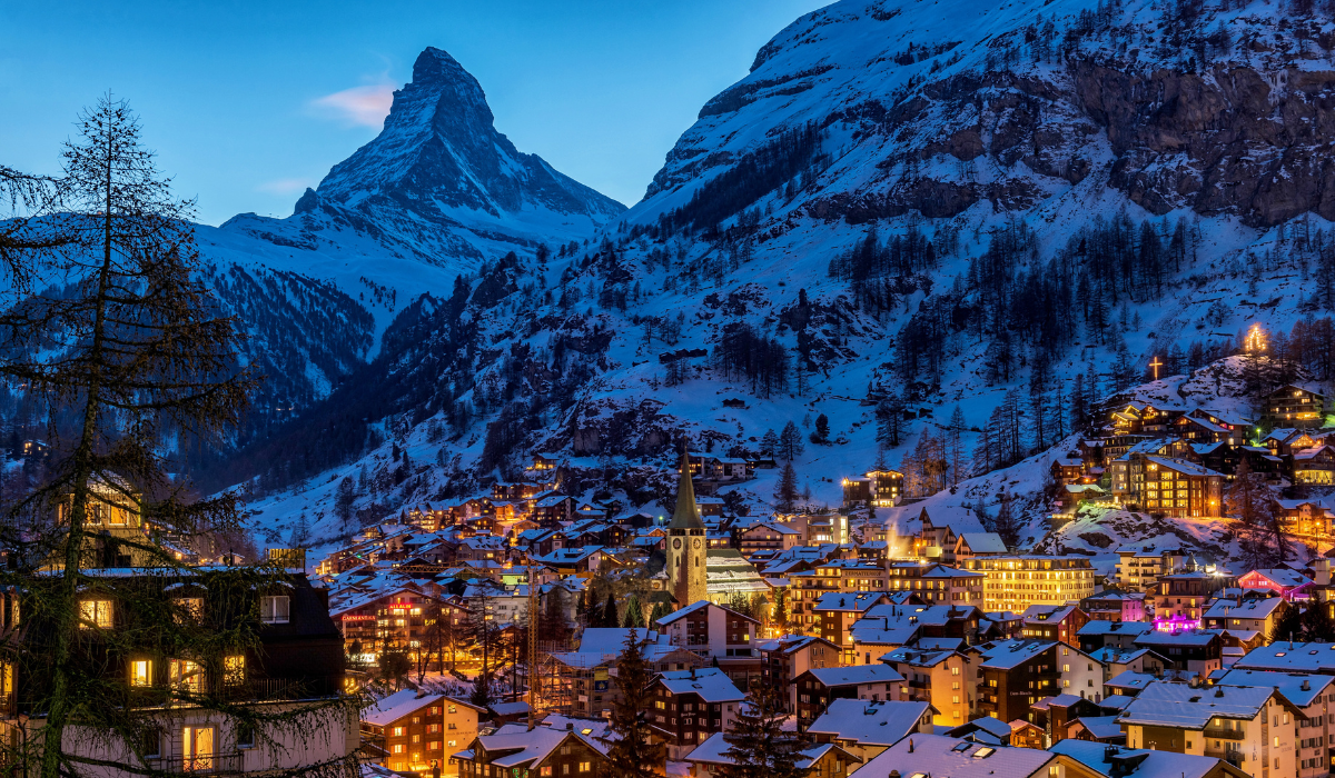twilight hour, view of the Zermatt village with building lights giving off warm golden glow. the matterhorn stands majestically in the background.