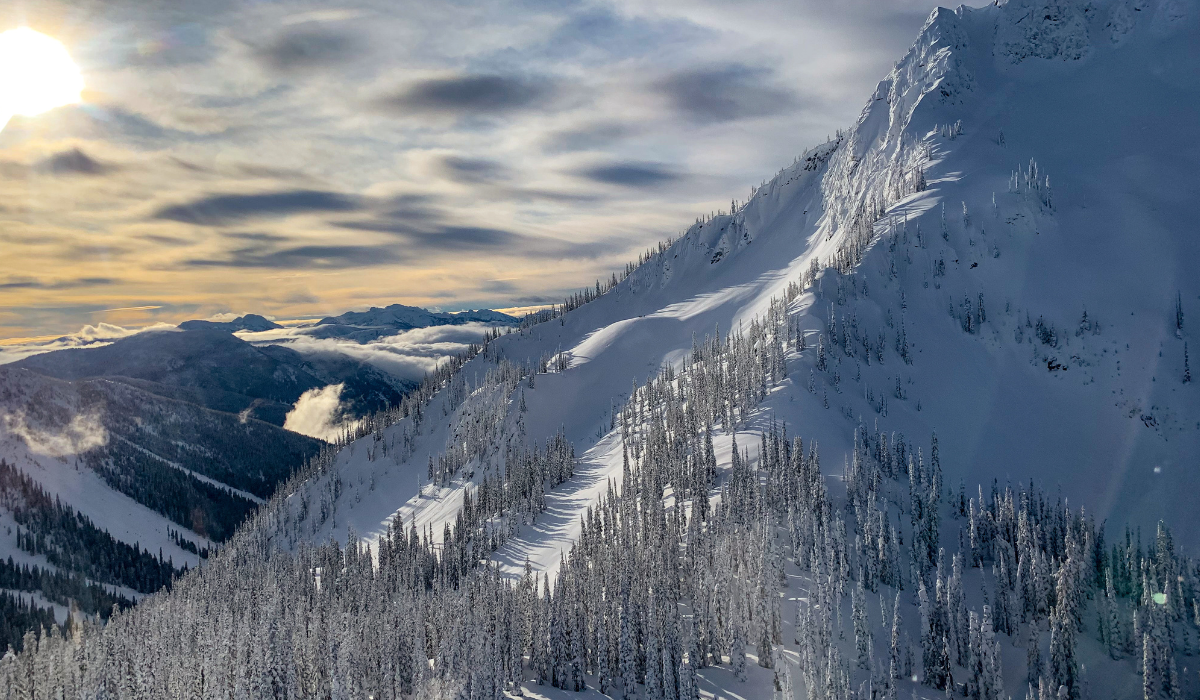 low sitting sun beams with large steep mountain covered in snow with white snow-covered alpine trees sitting at its lower levels.